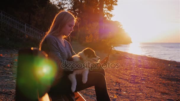 Genç kadın parkta bir göl ya da deniz kıyısında bir e-kitap okuma. Gün batımında, onu köpek yavrusu köpek kollarında bir bankta oturan — Stok video