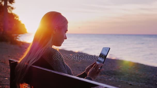Giovane donna con i capelli lunghi seduta su una panchina del parco. Utilizza tablet. Al tramonto visto un lago o il mare — Video Stock