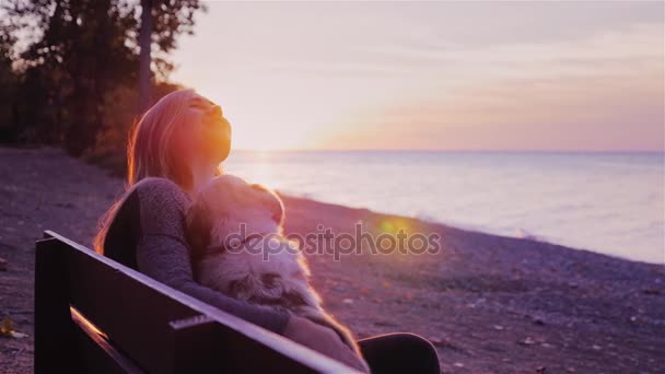 Attractive woman plays with her dog. Sitting on a bench in the park against the backdrop of a beautiful sunset over the sea — Stock Video