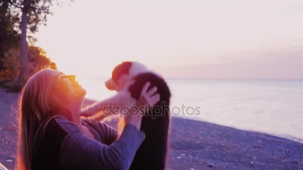Играй со своей любимой собакой. Женщина бросает твоего щенка на верх, играя с ним. На фоне заката моря или озера — стоковое видео