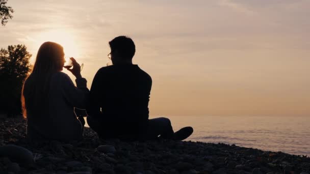 Silhouettes d'un jeune homme asiatique et d'une femme blanche. Asseyez-vous sur la plage au coucher du soleil, buvez du vin dans des verres — Video