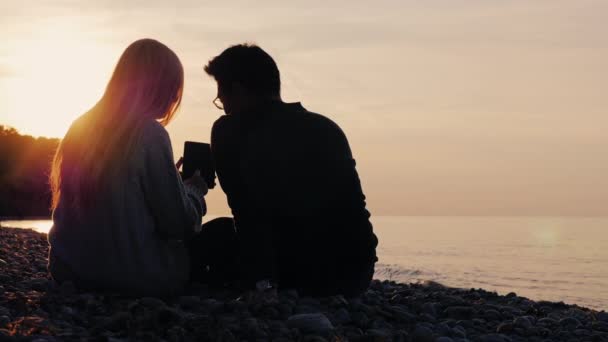Silhouetten von Mann und Frau, die am Kiesstrand sitzen und Tabletten genießen. bei Sonnenuntergang, dem Meer oder einem See. Rückansicht, multiethnisches Paar — Stockvideo
