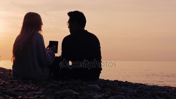 Tecnología en la naturaleza. Hombre asiático con gafas y una mujer tienen un descanso en la naturaleza, disfrutar de la tableta. Al atardecer cerca del mar o del lago — Vídeos de Stock