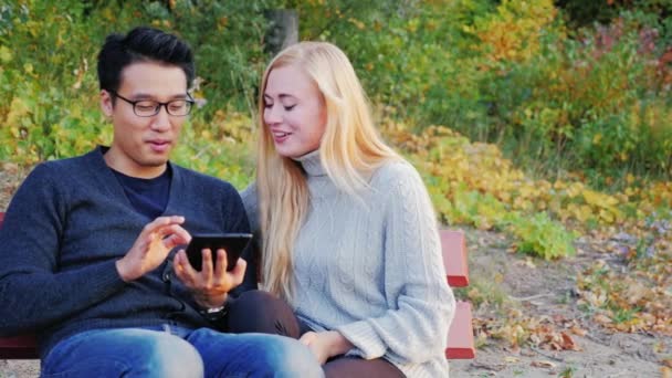 Молодой человек и женщина разговаривают в парке, наслаждайтесь планшетом. Кавказка и кореянка — стоковое видео