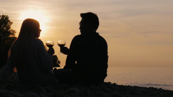Romantica coppia multietnica che beve vino sulla spiaggia o sul lago. Si possono vedere le loro sagome dietro al tramonto. Asiatico uomo e caucasico donna — Video Stock