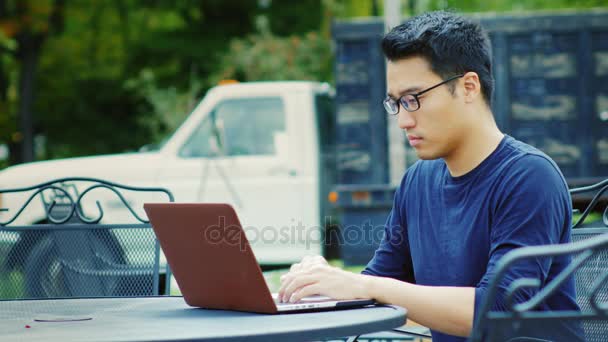 Aziatische man aan het werk met de laptop. Buiten op een achtergrond van oude vrachtwagen — Stockvideo