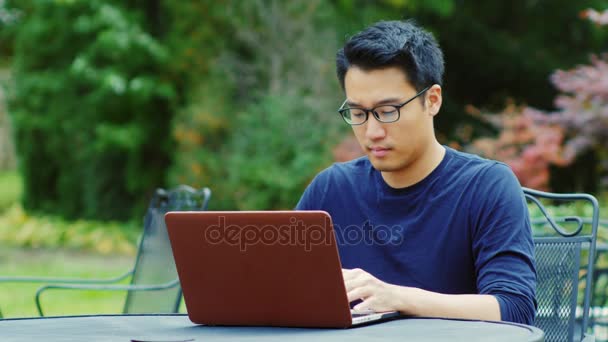 Hombre asiático con gafas funciona con un ordenador portátil. Sentado en la terraza de verano en los jardines o cafés . — Vídeos de Stock