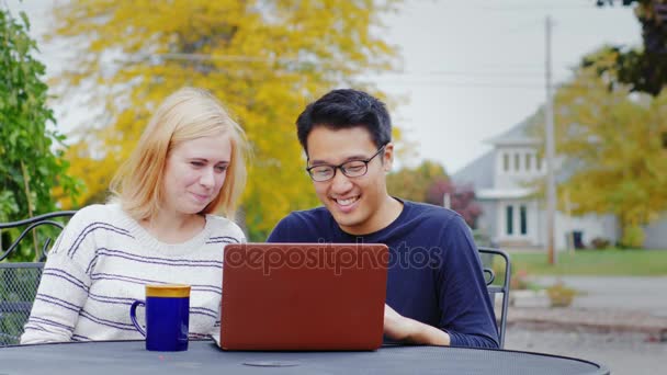 Dos amigos multiétnicos: un hombre coreano y una mujer caucásica trabajando juntos o viendo un video en una computadora portátil. Sonríe, ríe, diviértete. — Vídeos de Stock