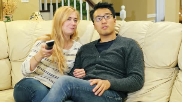 Przyjaciele oglądania telewizji w salonie. Koreański mężczyzny i kobiety rasy kaukaskiej. Trzymać w ręku pilota — Wideo stockowe