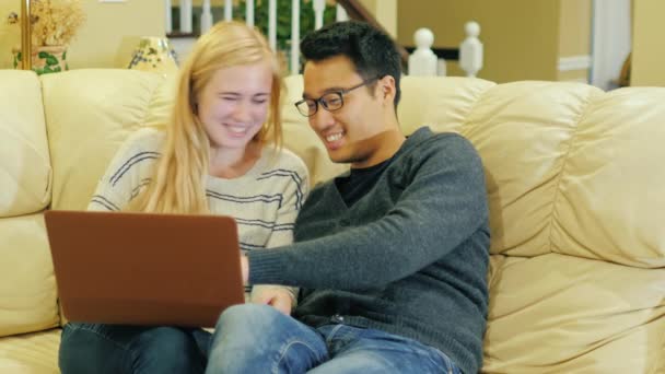 Multinacionális pár pihentető otthon, ül a kanapén, laptop használata — Stock videók