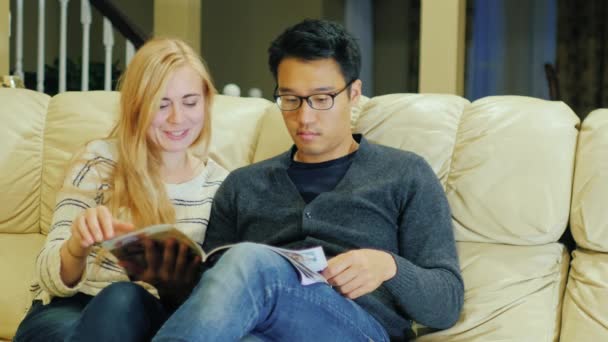 Μαζί, διαβάζοντας ένα περιοδικό. Νεαρό ζευγάρι πολυεθνικό ξοδεύει το χρόνο στο σπίτι. — Αρχείο Βίντεο