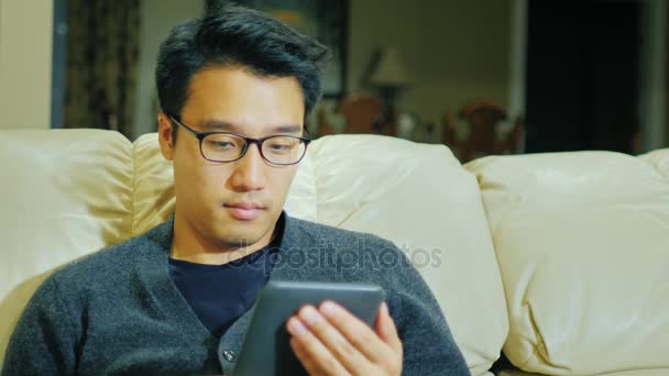 Un jeune Asiatique avec des lunettes. Il est assis sur le canapé à la maison, lisant e-book — Video