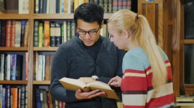 Asya adam gözlük ve beyaz kadının birlikte kütüphanede kitap izliyor. Rafları kitaplarla arka planı