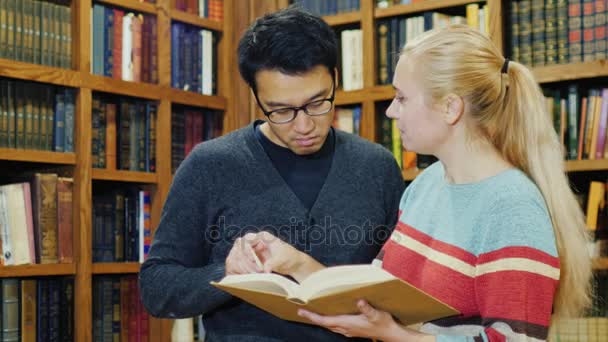 Homem coreano intilligentny e mulher caucasiana de pé juntos assistindo o livro na biblioteca. Estudando na Universidade — Vídeo de Stock