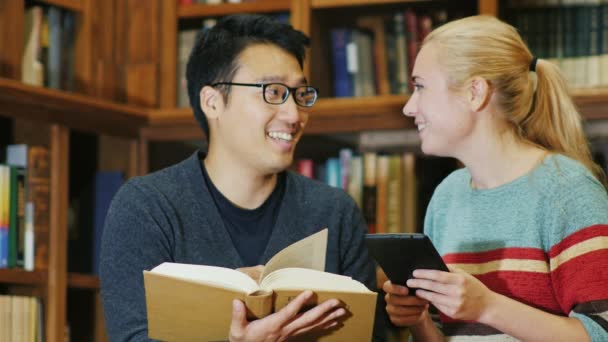 Hombre coreano sonriente hablando con una mujer en la biblioteca. disfrutar de la tableta — Vídeos de Stock