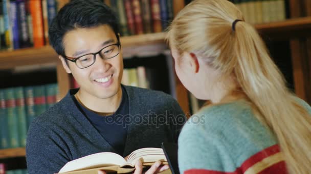 Studia w kolegium. Uśmiecha się kobieta oschaetsya asian mężczyzna w bibliotekę z książkami na tle półki — Wideo stockowe