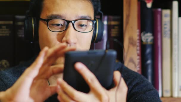 Portrét asijského muže v brýlích. Tablet využívá v knihovně. On sedí na pozadí regály. Měl na sobě sluchátka — Stock video