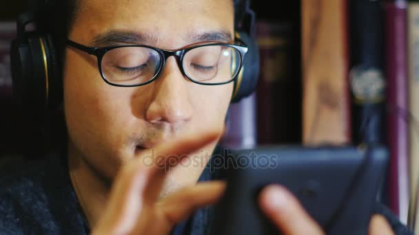 Ritratto di un coreano con gli occhiali. Sta usando un tablet digitale, ascoltando musica con le cuffie. Sullo sfondo di scaffali con libri — Video Stock