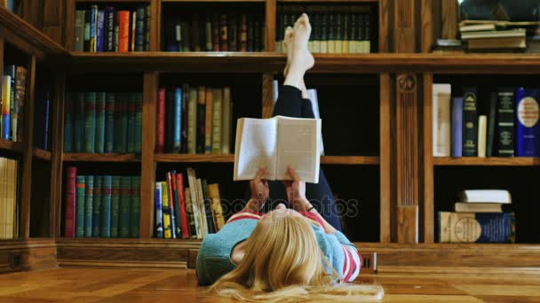 Fiatal nő fekve egy könyvet olvas. Hazudik a könyvtárban, a háttérben a polcokon könyvek — Stock videók