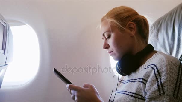 Joven mujer atractiva está volando en un avión. Utiliza un smartphone escuchando música — Vídeo de stock