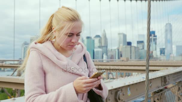 Mujer joven escribiendo un sms en su teléfono inteligente, de pie en la barandilla del puente de Brooklyn en Nueva York — Vídeo de stock