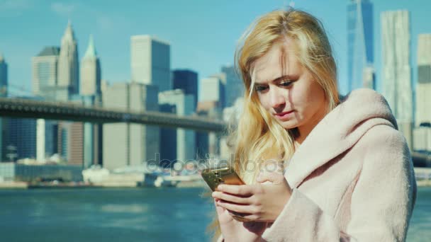 Nueva York, en el contexto de Manhattan y Brooklyn Bridge turista en un abrigo rosa escribiendo en su teléfono inteligente . — Vídeos de Stock