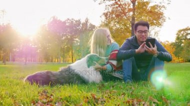 Genç çok etnik gruptan oluşan bir çift rahatlatıcı köpeğiyle parkta. Asya adam beyaz kadın köpekle oynamayı bir tablet bakan