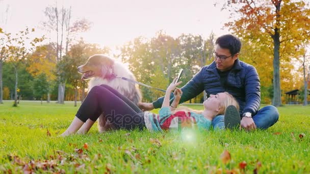 Interracial fiatal pár pihenő parkban egy kutya. Simogatta a kutya, a kaukázusi nő olvasó e betű-könyv olvasó vagy tabletta ázsiai ember használ — Stock videók