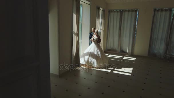 Жених и невеста в элегантных платьях стоят у окна в большом зале — стоковое видео