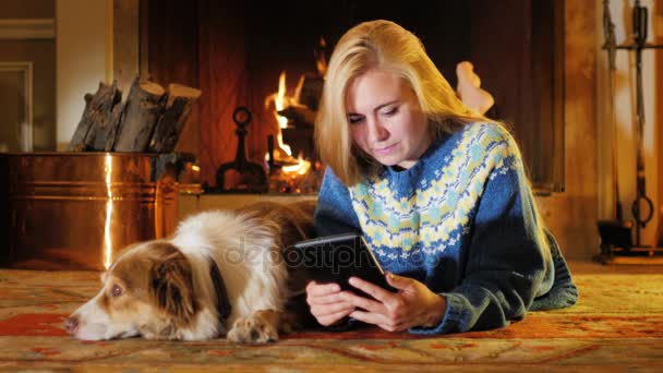 Eine junge attraktive Frau genießt die Tablette. in einem komfortablen Haus auf einem Hintergrund des Feuers. Daneben steht ein Hund — Stockvideo