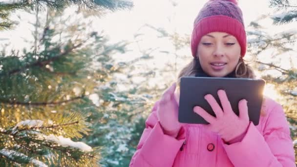 Mujer joven en ropa de invierno rosa disfruta de la tableta. Ella está en un bosque de invierno, detrás del sol brilla maravillosamente — Vídeos de Stock