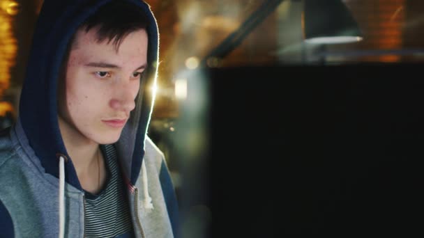 Κουρασμένο εφηβικό αγόρι που κάθεται μπροστά από οθόνες υπολογιστών αργά το βράδυ. Φοράει κουκούλα — Αρχείο Βίντεο
