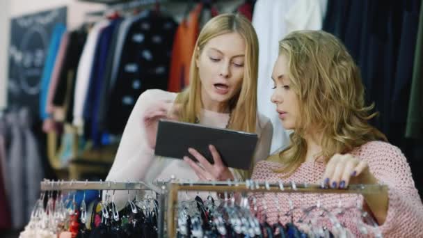 Dos mujeres jóvenes hacen compras en una tienda de ropa. Disfruta de tu tablet, mira cosas en el catálogo y en los stands de la tienda — Vídeos de Stock