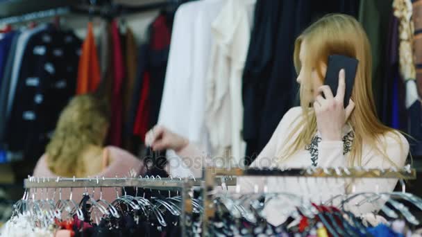 Хороший шопинг и общение по телефону. Молодая женщина выбирает вещи в магазине женской одежды — стоковое видео
