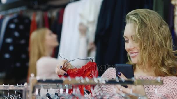 A(z) store női ruházat. Egy nő néz egy fényképet a telefont, és vedd fel a vásárlás, a háttérben a vevő van is látszó ruhát — Stock videók