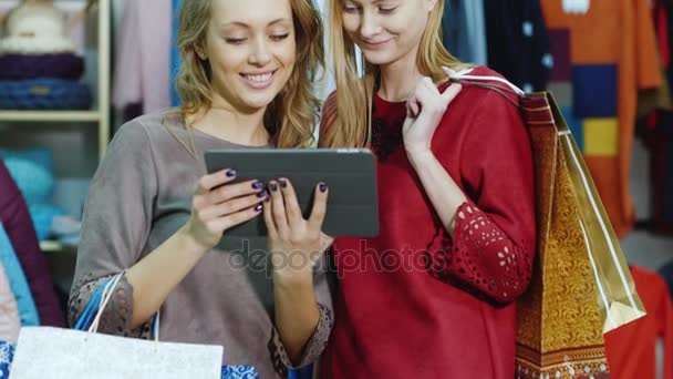 Duas mulheres elegantes em vestidos com sacos de compras. Em pé na loja: aproveite o seu tablet, sorrindo. Compras bem sucedidas — Vídeo de Stock