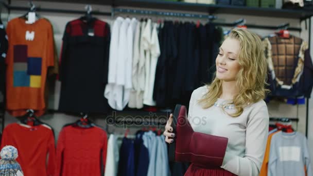 An attractive young woman chooses warm winter shoes in a clothing store. Smiles: looking at the boots red — Stock Video