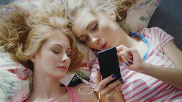 Duas jovens deitam-se de frente para a cama. Desfrute de smartphones, ouça música. Vista superior, belo cabelo deitado em um travesseiro — Vídeo de Stock