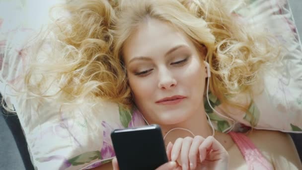 Портрет привлекательной молодой женщины, лежащей на кровати, использует смартфон, слушая музыку. Вид сверху — стоковое видео