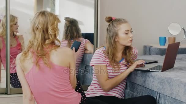 Две молодые женщины отдыхают дома, пользуются разными устройствами, телефоном, ноутбуком. Девушки с пижамной вечеринки — стоковое видео