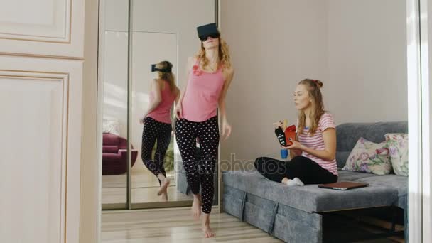 Due amiche si riposano a casa. Uno balla nel casco della realtà virtuale, l'altro mangia la loro scatola di carta alimentare. Pigiama partito amici — Video Stock