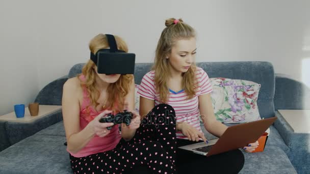 Двое друзей развлекаются дома. Играя в шлеме виртуальной реальности, используйте ноутбук. Девушки с пижамной вечеринки — стоковое видео