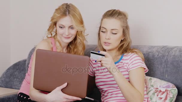 两个朋友进行在线采购。年轻的女人坐在沙发上，一台笔记本电脑的手里拿着一张信用卡 — 图库视频影像