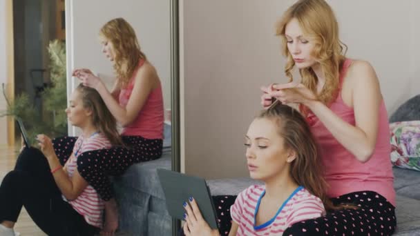 Dos mujeres jóvenes relajándose en casa, disfrutando de la tableta, cabello trenzado. Pijama partido novias — Vídeo de stock