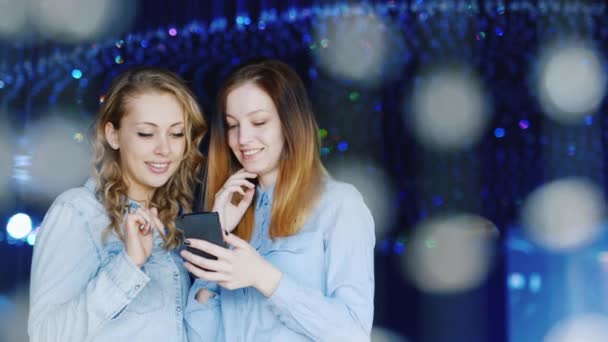 Twee aantrekkelijke jonge vrouwen genieten van een smartphone in een nachtclub. Ze staan rond van glazen ornamenten — Stockvideo