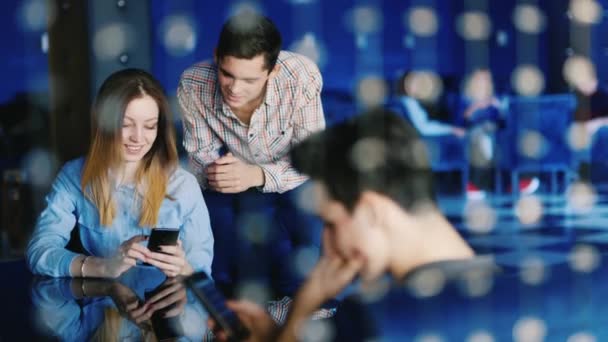 En grupp unga människor vilar på ett café. Ett ungt par söker något på skärmen smartphone, i förgrunden mannen använder en surfplatta — Stockvideo