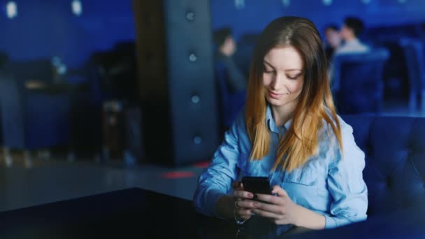 Genç bir kadın yalnız bir kafe ya da gece kulübü oturur, bir telefon kullanır — Stok video