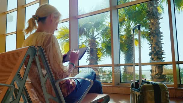 Una giovane donna gode di un tablet nel terminal dell'aeroporto. Giorno soleggiato, fuori dalla finestra crescono palme. Ritorno dal resort — Video Stock