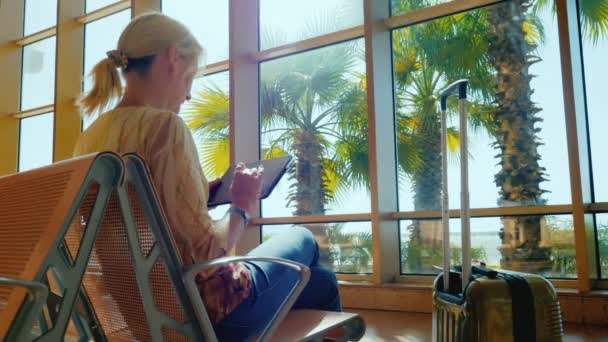 Una joven disfruta de una tableta en la terminal del aeropuerto. Día soleado, fuera de la ventana crecen palmeras. Regreso del resort — Vídeo de stock