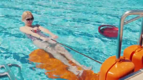 Jovem está se exercitando em um simulador na piscina. Descanso ativo em ar fresco, atividade no resort — Vídeo de Stock